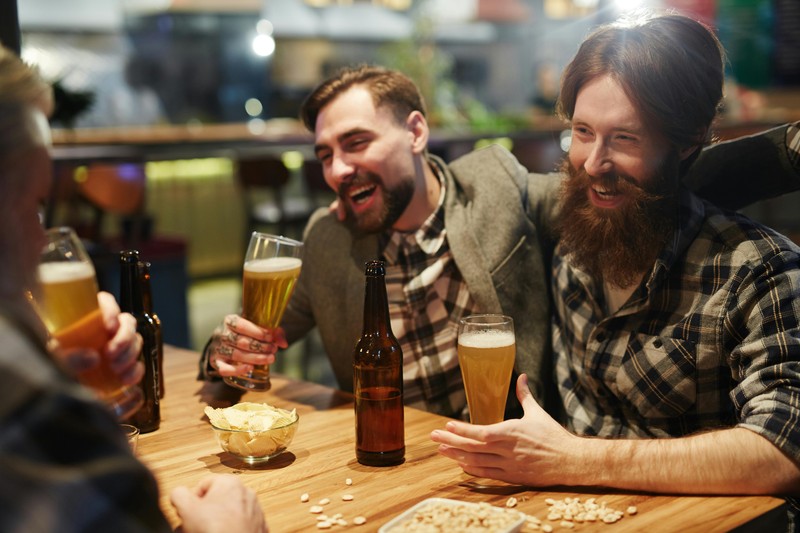 A group of friends laughing together around a wooden table in a cabin with drinks and a rustic setting.