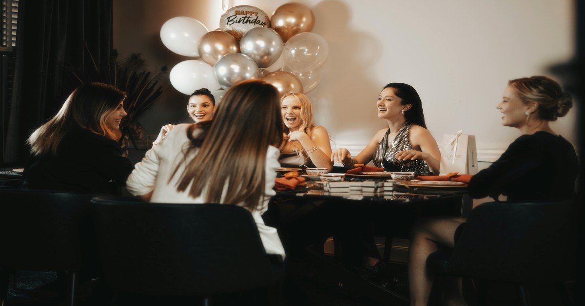 A group of women laughing and planning a bachelorette party around a table with decorations and drinks